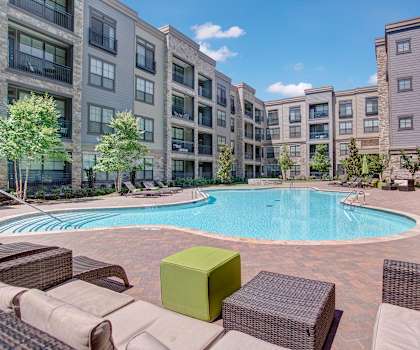 The large, turquoise outdoor pool with green pool umbrellas, tan pool chairs with greenery, trees, and the apartments in the background at the Enclave at Brookside Apartment Homes. 