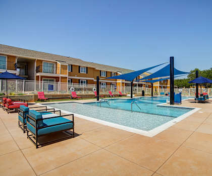 The turquoise outdoor pool with pool chairs, tables, and chairs with the apartments in the background at Indigo Apartments. 