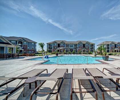 The turquoise outdoor pool with pool chairs, tables, and chairs with the apartments in the background at Liberty Pointe Apartment Homes. 