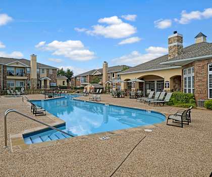 The turquoise outdoor pool with pool chairs, tables, and chairs with the apartments in the background at Quail Landing Apartment Homes. 