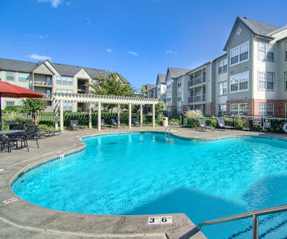 The large, turquoise outdoor pool with red umbrellas, pool chairs, tables, and chairs with the light blue apartments in the background at Villas at Preston Lakes Apartment Homes. 