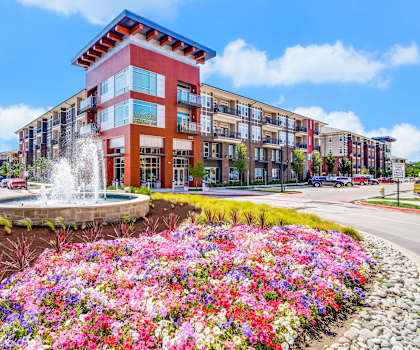 The exterior of Harmony Luxury Apartments, showing a beige, red, and brown apartment building with a fountain and flower beds in front. 