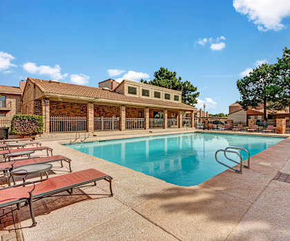 The turquoise outdoor pool with pool chairs, tables, and chairs with the apartments in the background at Las Colinas Apartments.