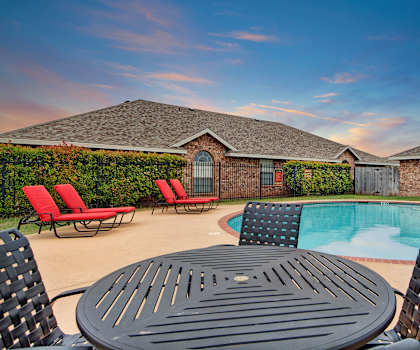 The turquoise outdoor pool with pool chairs, tables, and chairs with the apartments in the background at Lexington Court Apartment Homes. 