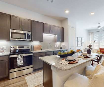 The living room with a couch, plush chairs, coffee table, decorations, a TV, and view of the dining room and the kitchen with marble counters and black appliances at Magnolia on the Green Apartments. 