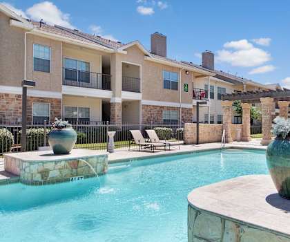 The turquoise outdoor pool with pool chairs, tables, and chairs with the apartments in the background at Mission Green Apartments. 