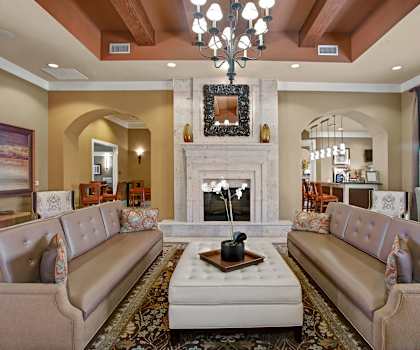 The amazing resident lounge with a fireplace, a mirror above it, tan couches with an orchid, decorations, large windows with curtains, white plush chairs, a chandelier, and white coffee tables at the Palms at Briarwood Apartment Homes. 