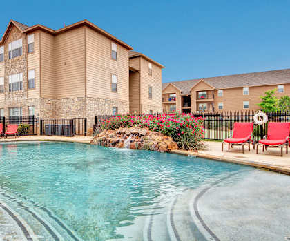The large, turquoise outdoor pool with a small, rocky waterfall, red pool chairs with greenery, trees, and the apartments in the background at the Reserve at Abilene Apartment Homes. 