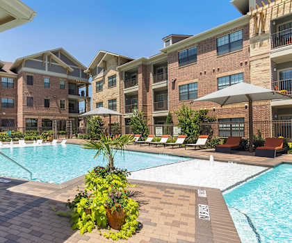 The large, turquoise outdoor pool with a fountain surrounded by palm trees, pool chairs, tables, cabanas, and chairs with the apartments in the background at Sorrel Fairview Apartment Homes. 