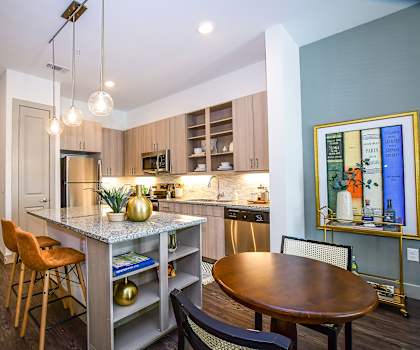 An artsy kitchen with modern lighting, a kitchen island with two burnt orange bar chairs, light gray spacious cabinets, granite countertops, kitchen utensils, and wall art with a dining table and two chairs at the Crosby at the Brickyard Apartment Homes. 