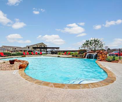 The large, turquoise outdoor pool with a fun fountain with rocks, red pool chairs with greenery, trees, and the apartments in the background at the Residence at Heritage Park Apartment Homes. 