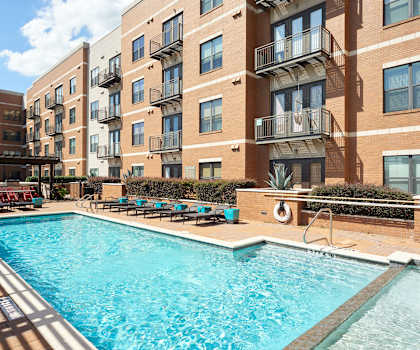 The large, turquoise outdoor pool with pool chairs, tables, and chairs with the apartments in the background at the Uptown Apartment Homes. 