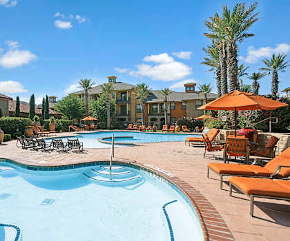 The two large, turquoise outdoor pools with orange pool chairs, orange umbrellas, tables, and chairs with the apartments in the background at Tuscany at Faundree Apartment Homes. 