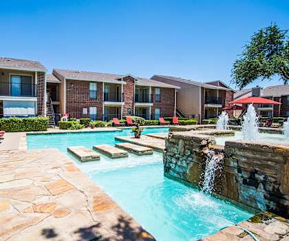 The large, turquoise outdoor pool with fountains and steps in the pool to the other side of the pool with pool chairs, tables, and chairs with the apartments in the background at Wildflower Apartment Homes. 