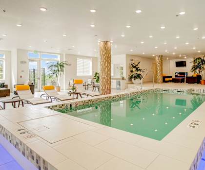 The indoor pool with tables, chairs, pool chairs, and palms trees, TV, and a fireplace in the background at Heritage Hills. 