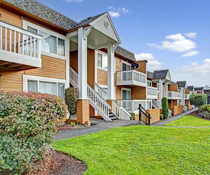 The beautiful exterior of the tan and white apartment buildings and the green space with grass, trees, and shrubs at Whispering Cedars Apartment Homes. 