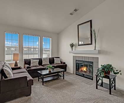The spacious living room with a dark velvet couch, gray throw pillows, large windows, a ceiling fan, wall art, dark velvet plush chair, decorative plants, coffee table, decorations, large windows, a TV, and glass doors to the balcony at St. James Place Apartment Homes. 