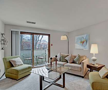 The spacious living room with a light pink couch, throw pillows, large windows, wall art, two green plush chairs, coffee table, decorations, large windows, a TV, and glass doors to the balcony at Sunset Ridge Apartment Homes. 