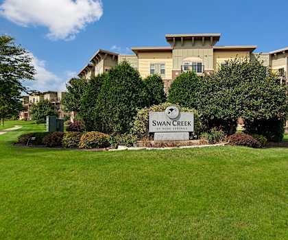 The silver main sign in front of the apartments surrounded by green grass, shrubs, and trees at Swan Creek Apartment Homes. 