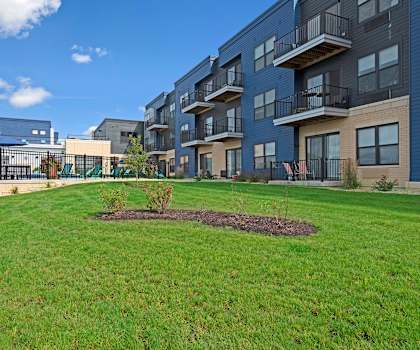 The exterior green area with grass, plants, and tress with the blue apartment buildings and the pool area in the background. 