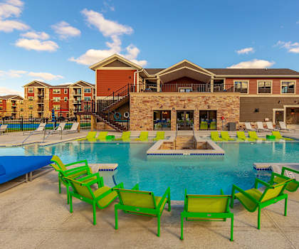 The large, turquoise outdoor pool with neon green, white, and blue pool chairs, tables, and chairs with the apartments in the background at the Park Apartment Homes. 