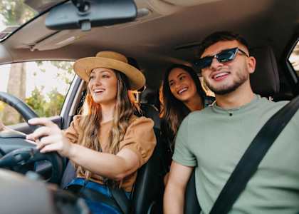 Three people are sitting in a car, with the woman driving, one woman in the back seat, and one man in the passenger seat