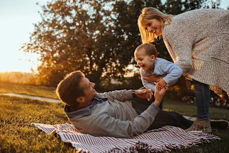 A family of three is sitting on a blanket in a park during sunset.
