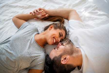 A couple is lying on a bed, holding hands and smiling at each other.