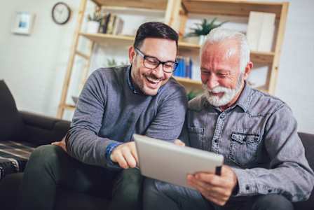 Two men are sitting on a couch, one holding a tablet and both smiling.