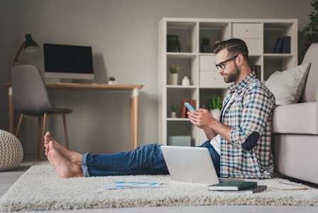 A man is sitting on the floor with his feet up, using a laptop and holding a smartphone.