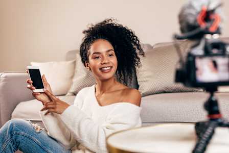 A woman is sitting on a couch holding a phone and recording a video.
