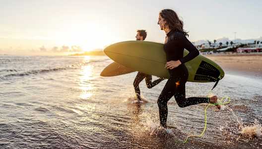 A person in a black wetsuit is running on the beach with a green surfboard and a yellow leash.