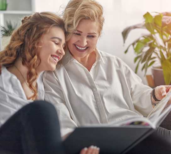 Two women are sitting on a couch, one is reading a book and the other is laughing.