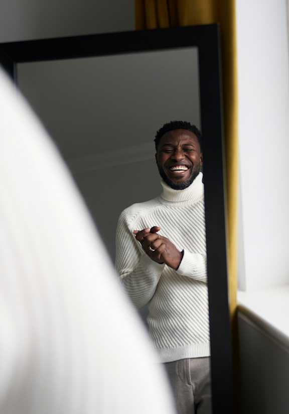 A man in a white sweater is looking at his reflection in a mirror.