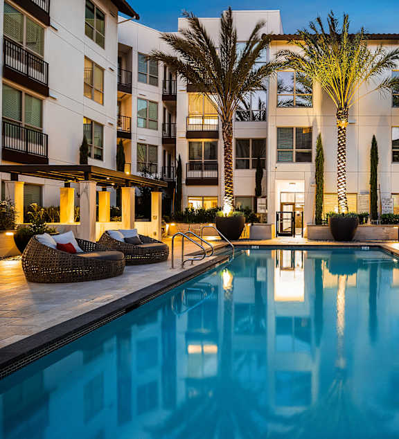 Twilight poolside view with glowing water, lush landscaping, and stylish lounge seating for relaxation
