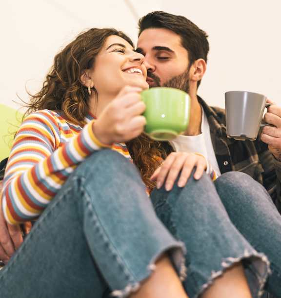 A man and woman are sitting on a couch, each holding a mug, and appear to be enjoying a moment together.