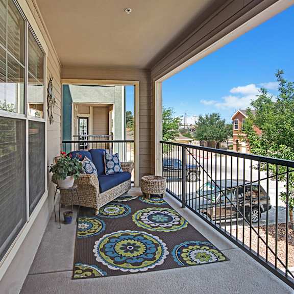 A concrete and metal balcony with a wicker couch and footrest with blue cushions and blue throw pillows, a potted plant with flowers, and a brown patterned rug with a gorgeous view of the other apartments and the parking lot at Palms at Briarwood Apartment Homes. 