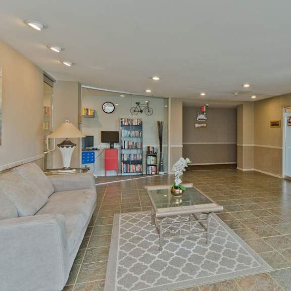 The lobby with a tan couch, glass coffee table with an orchid, a lamp on a coffee table, and an entertainment corner with a bookshelf and computer monitor at Somerset Pointe Apartment Homes. 