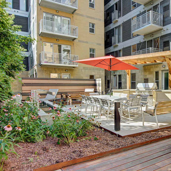 A patio with a grill area, a couch, and tables and chairs with an orange umbrella surrounded by the apartment buildings at Vue Apartment Homes. 