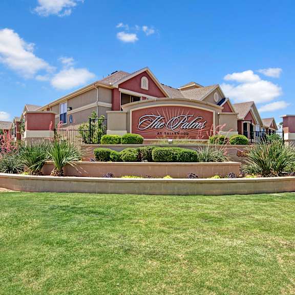 The exterior burnt orange sign with trees and surrounded by greenery, trees, and flowers with the buildings in the background of the Palms at Briarwood Apartment Homes. 