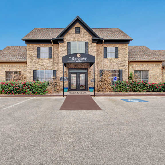 The exterior of the stone brick front office at Reserve at Abilene with flower bushes and the parking lot outside. 