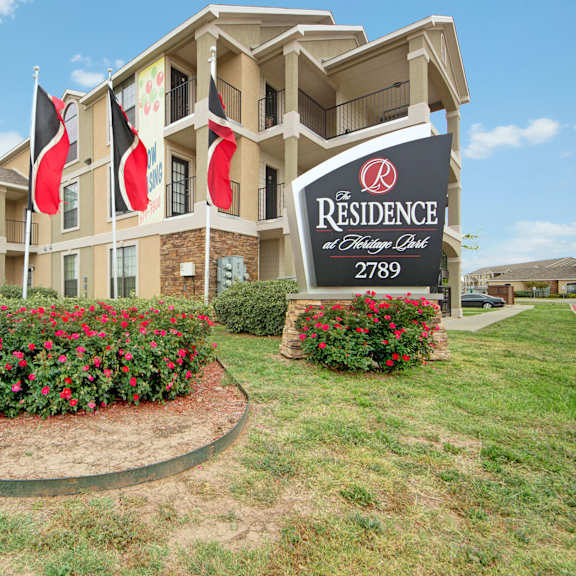 The black entrance sign, black and red flags, flower beds, and the front of the building of Residence at Heritage Park Apartment Homes. 
