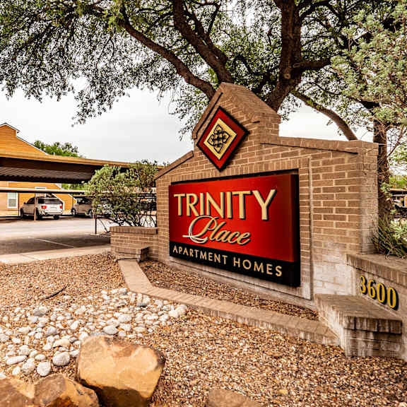 The exterior sign surrounded by rocks and a large tree behind it with the buildings in the background of Trinity Place Apartment Homes. 