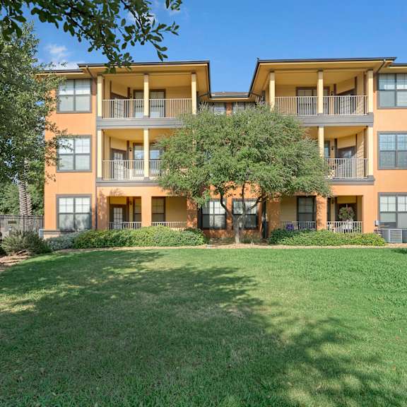 The beautiful, orange exterior of the apartments and the green space with grass, trees, and shrubs at Tuscany at Faudree Apartment Homes. 