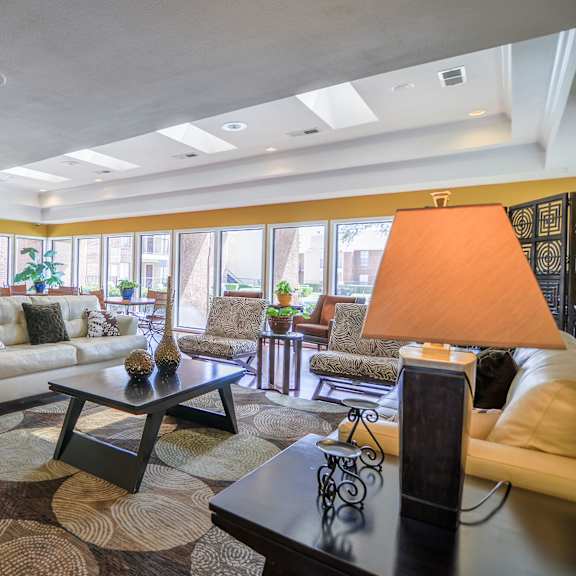 The resident lounge with couches, plush chairs, house plants, lamps, floor to ceiling windows showing the poor area, and coffee tables at Wildflower Apartment Homes. 