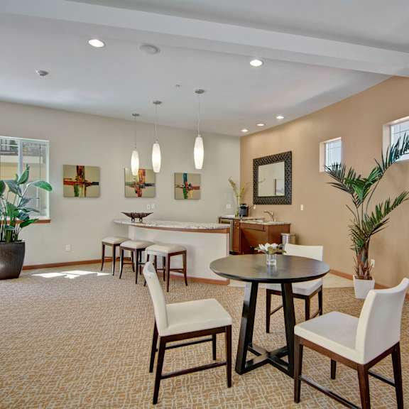 A resident lounge with a coffee bar, sink, refrigerator, and barstools, with a table, chairs, wall art, and plants at Ondine at Juanita Bay Apartments. 