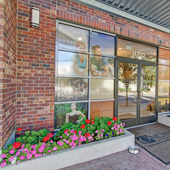 The entrance to Ondine at Juanita Bay Apartments with images of families and residents enjoying the apartments on the windows right by the doors, flower beds, and a door mat surrounded by brick at Ondine at Juanita Bay Apartments. 