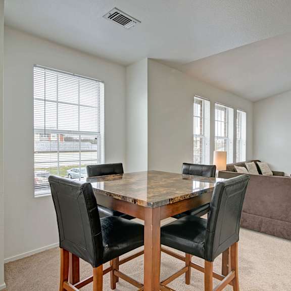 The dining room with a marble and wood table, four black leather dining room chairs, with a view of the living room with a brown couch, mirror, and fireplace at St. James Place Apartment Homes. 