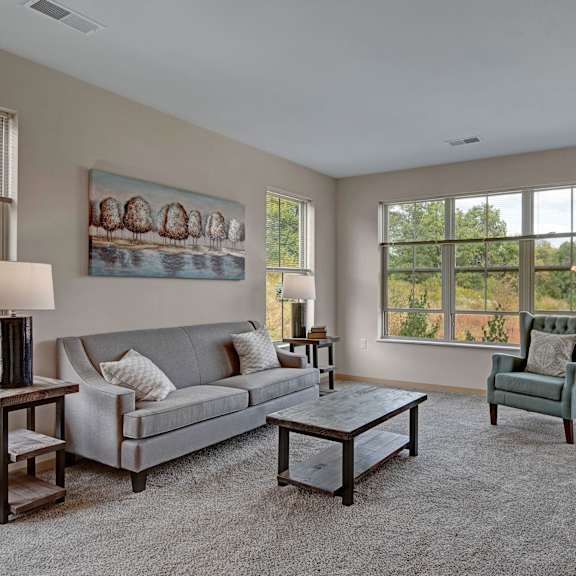 The spacious living room with a gray couch, white throw pillows, wall art, large windows, a ceiling fan, wall art, plush chairs, coffee table, decorations, large windows, a TV, and glass doors to the balcony at The Vue at Pinnacle Park Apartment Homes. 