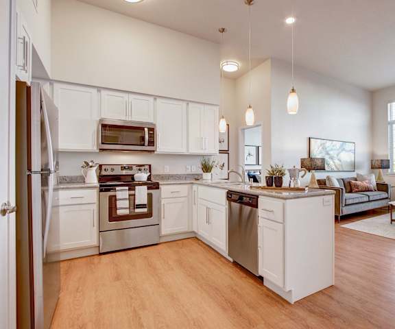 A large, modern kitchen with stainless steel appliances, decorative plants, bar counter with barstools, kitchen utensils, marble countertops with lots of room, unique lighting displays, a view of the living room, and large wooden cabinets at the Ritz Classic Apartment Homes. 